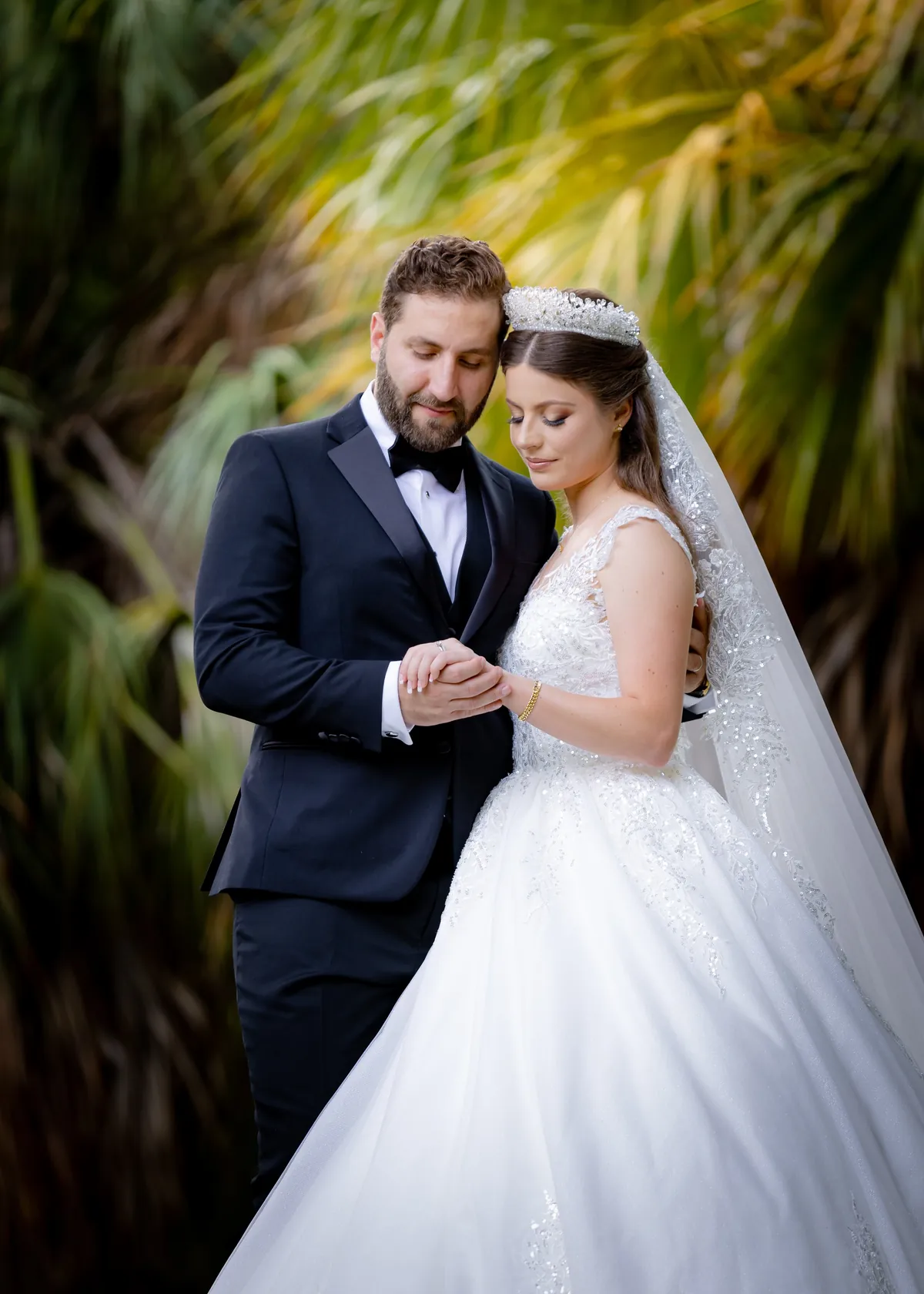 Bride in a white gown and tiara with the groom in a black tuxedo, palms behind them