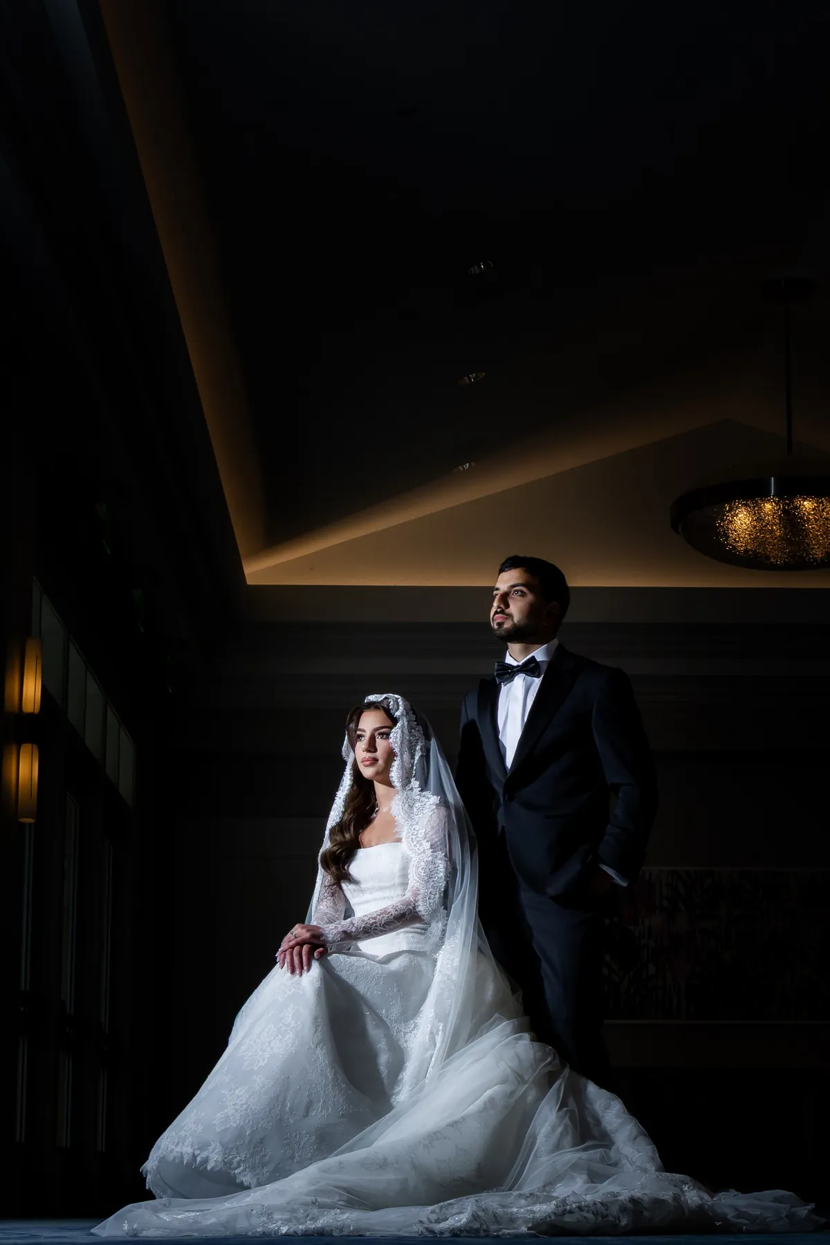 Seated bride and standing groom in a dark, dramatic reception hall