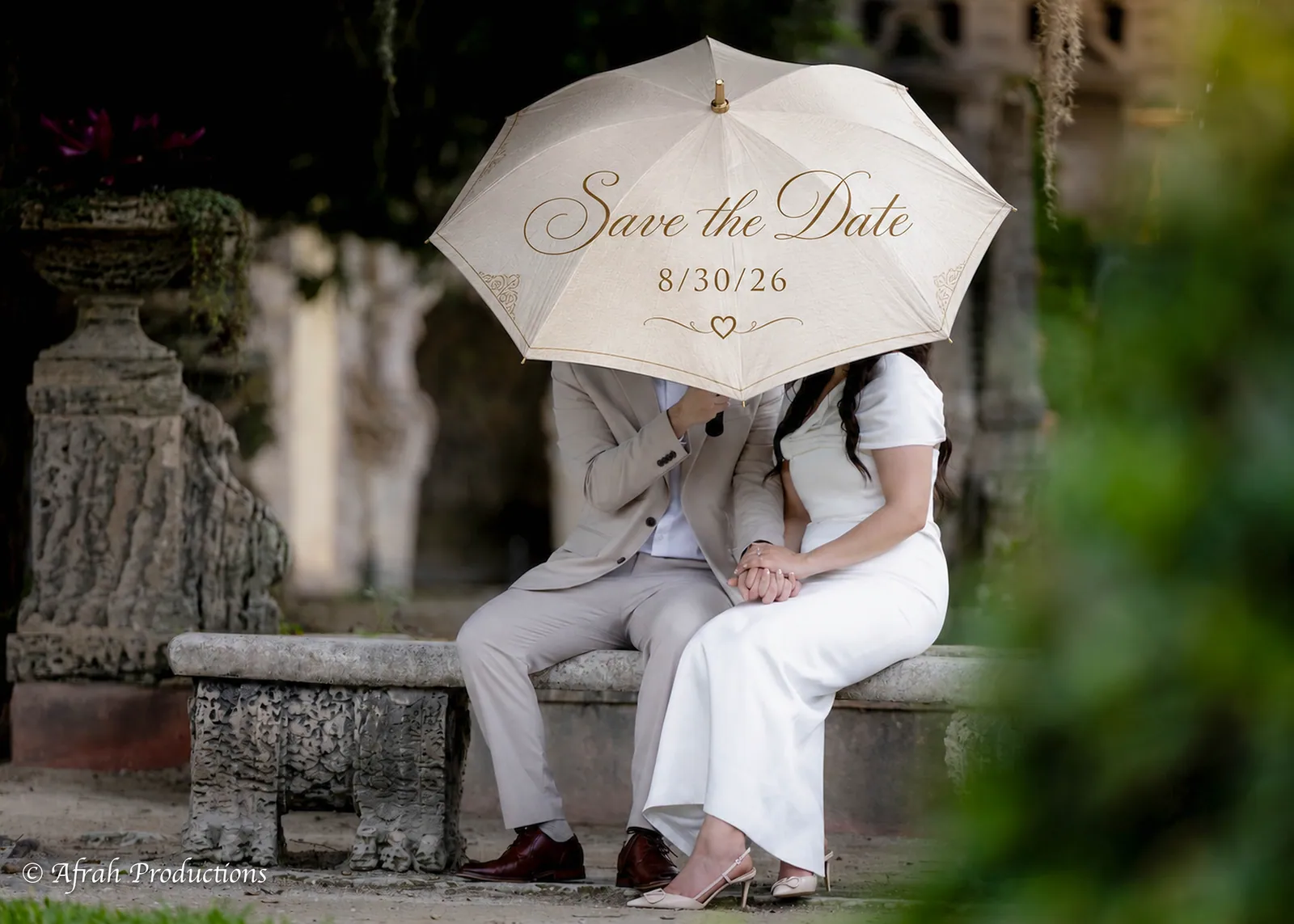 Couple under a Save the Date parasol on a stone bench in a garden