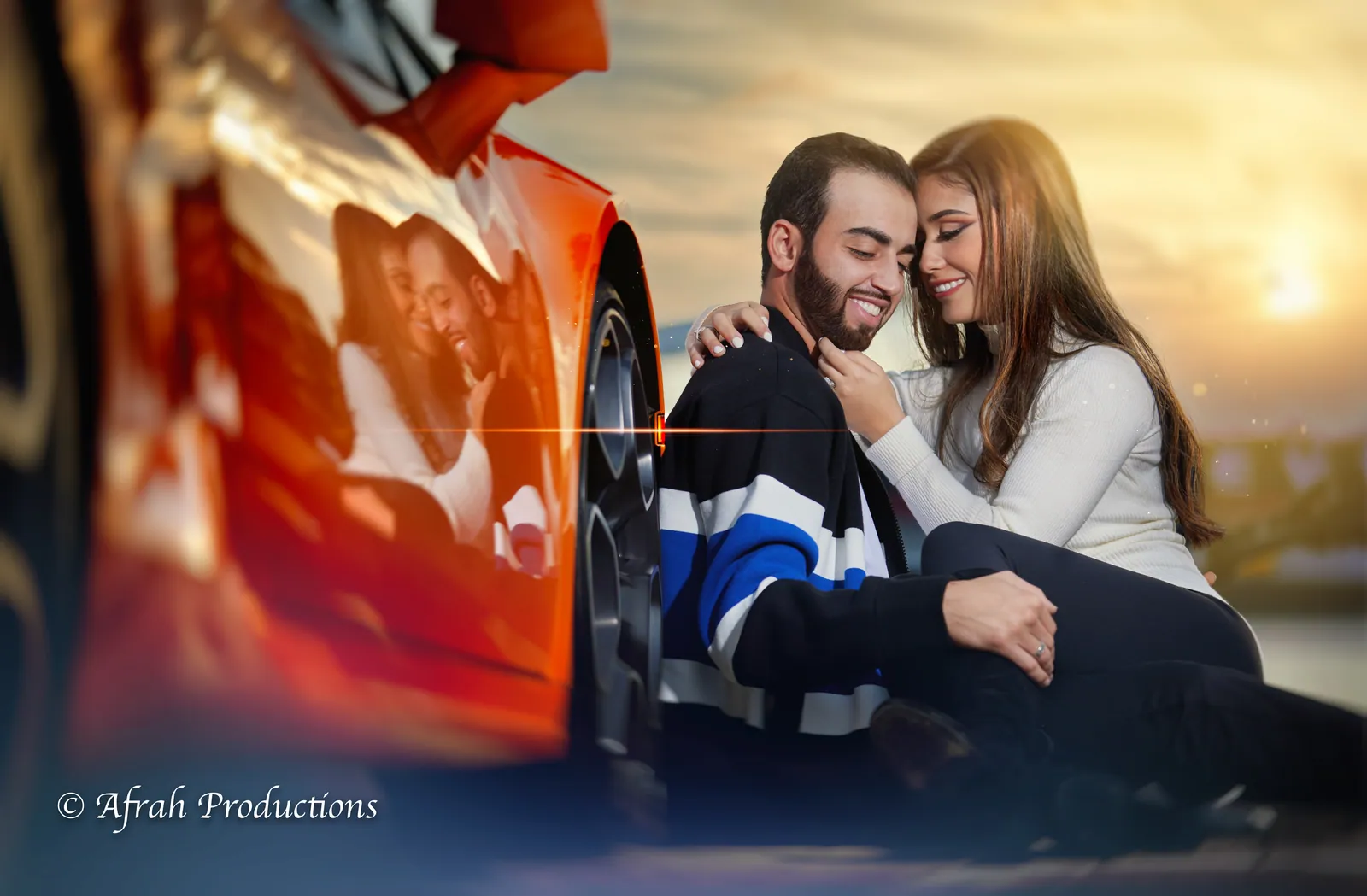 Couple seated beside a red sports car at sunset