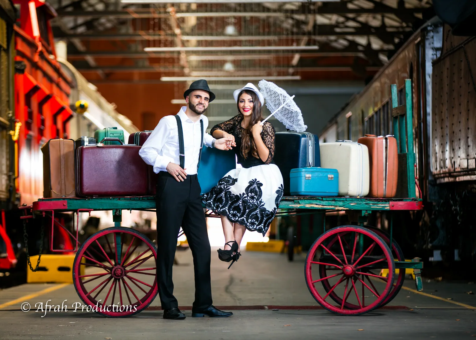 Couple on a vintage luggage cart in a train yard, parasol and lace