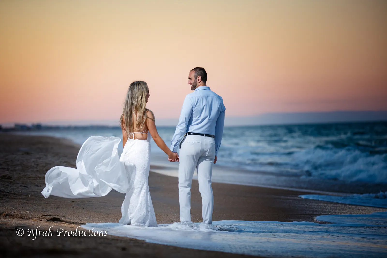 Couple at the water's edge at sunset, her dress lifting in the wind