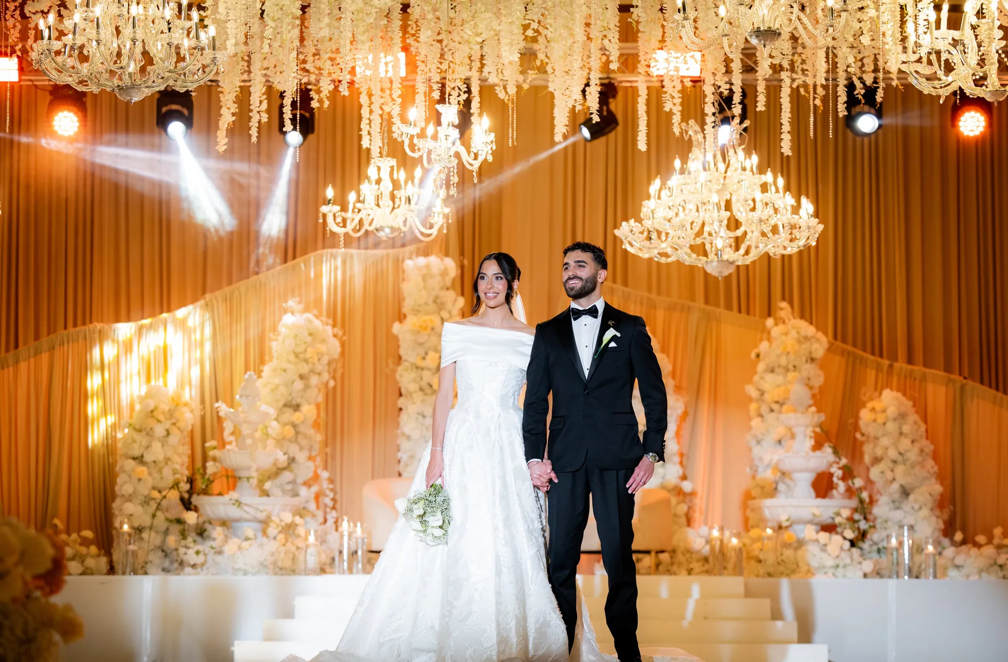 Bride and groom on stage at a reception with gold drapes and crystal chandeliers