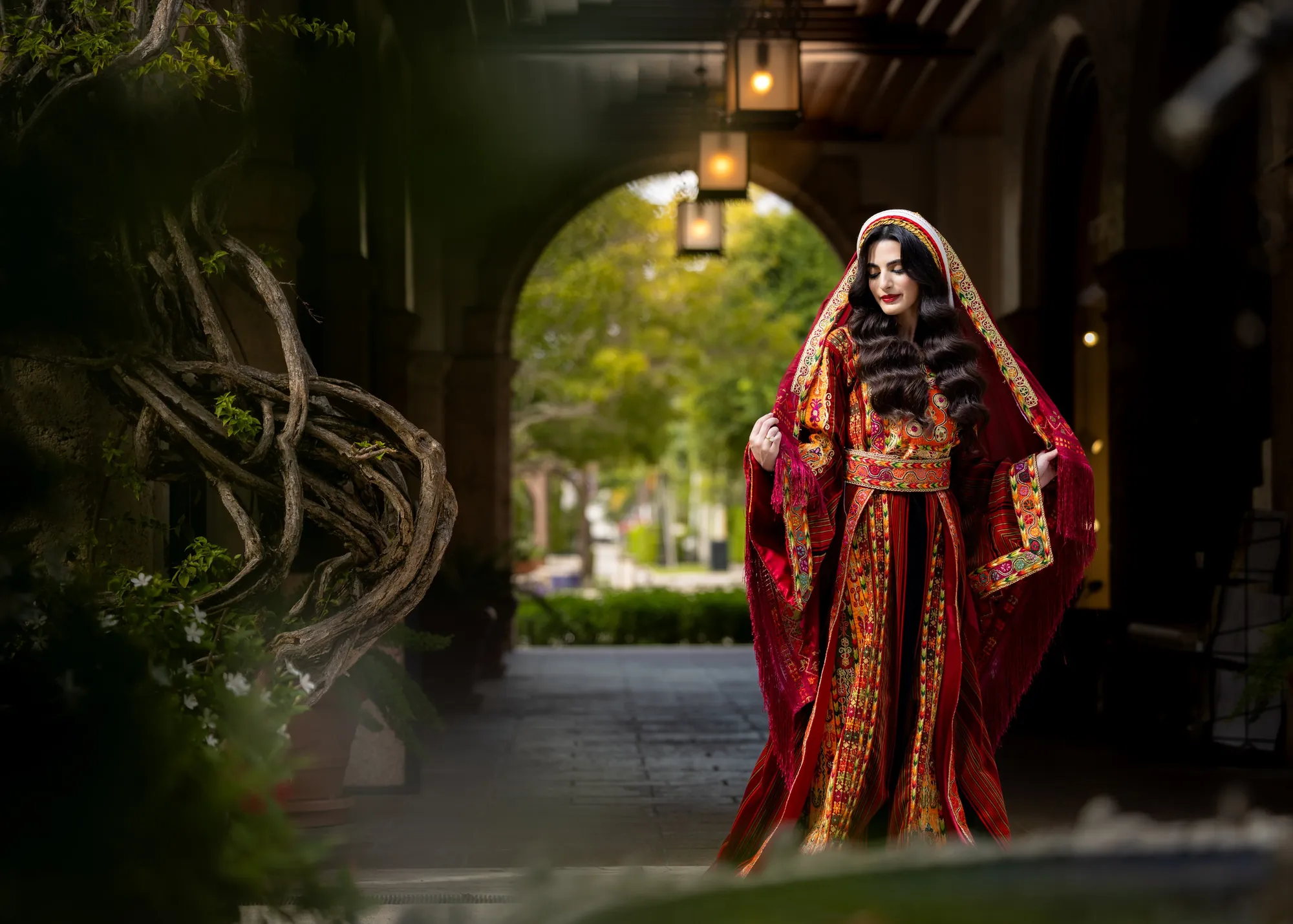 Bride in a red and gold embroidered traditional dress and veil walking through a stone arch