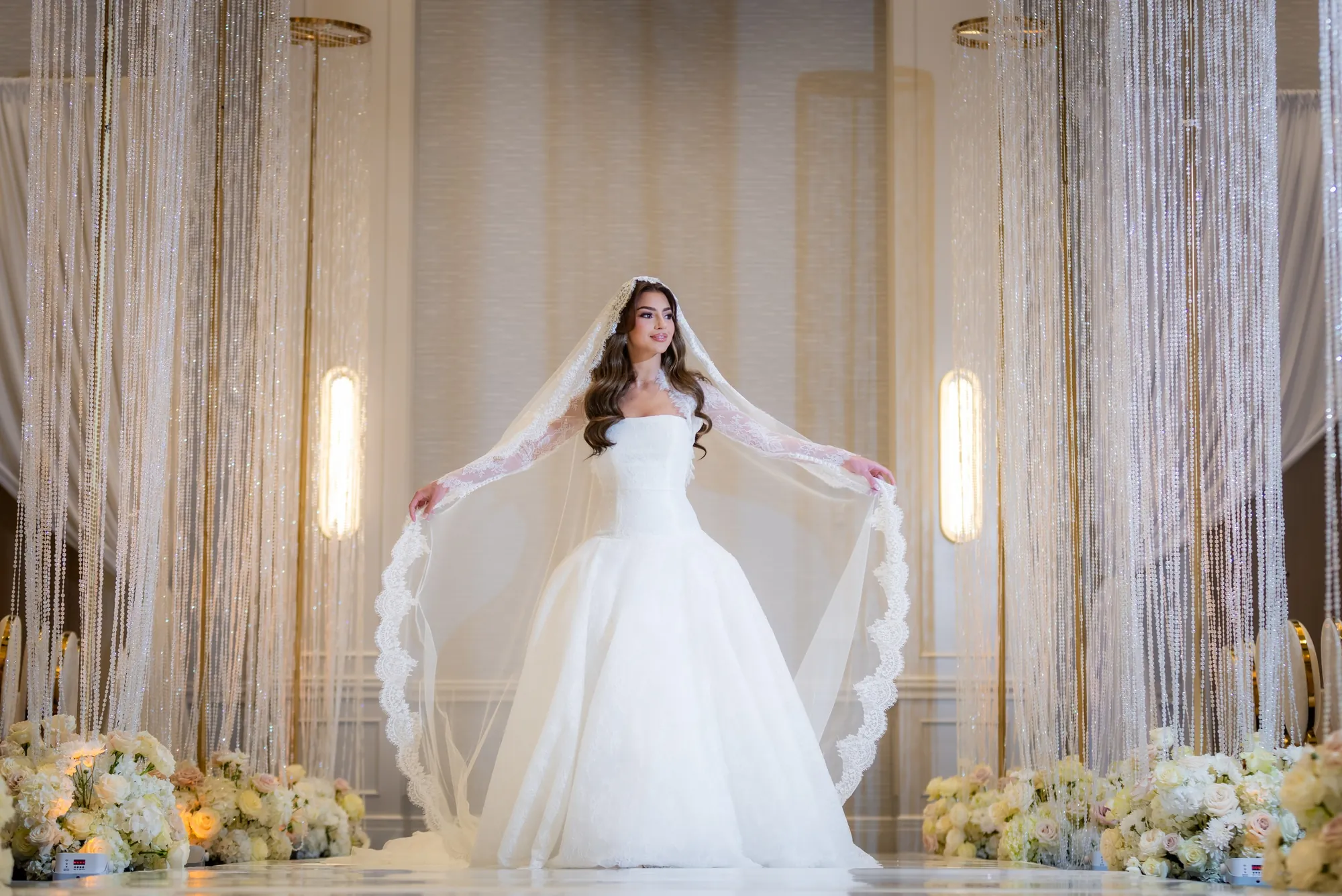 Bride holding her lace veil open between crystal bead curtains and white flowers